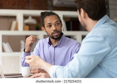 Multi ethnic diverse colleagues discuss collaborative task. African and Caucasian male workmates talking sit at workplace desk share ideas, skills, develop strategy solve business at meeting in office