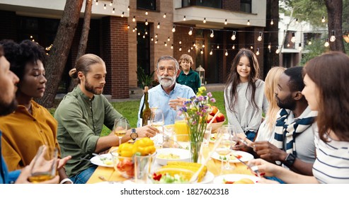 Multi Ethnic Family Sitting At Table With Meal Outdoor At Picnic And Senior Man Saying Toast. Mixed-races Happy Young And Old People Having Dinner And Toasting At Party Barbrque Celebration On Weekend