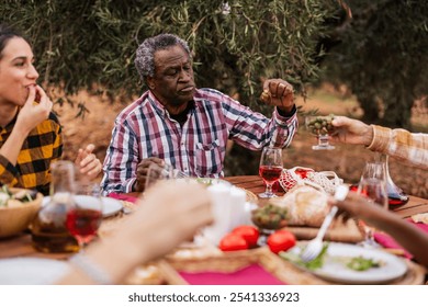 Multi ethnic farmers enjoying a healthy lunch with olives, bread, tomatoes and wine, sitting at a wooden table in an olive grove, tasting the fruits of their labor - Powered by Shutterstock