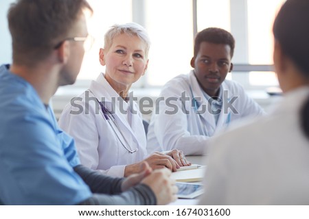 Multi ethnic group of doctors sitting at table, focus on mature woman heading committee meeting at medical council