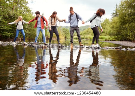 Multi ethnic group of five adult friends hold hands and help each other while carefully crossing a stream standing on stones during a hike