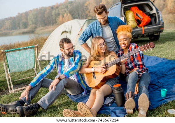 Multi ethnic group of friends dressed\
casually having fun playing guitar during the outdoor recreation\
with tent, car and hiking equipment near the\
lake