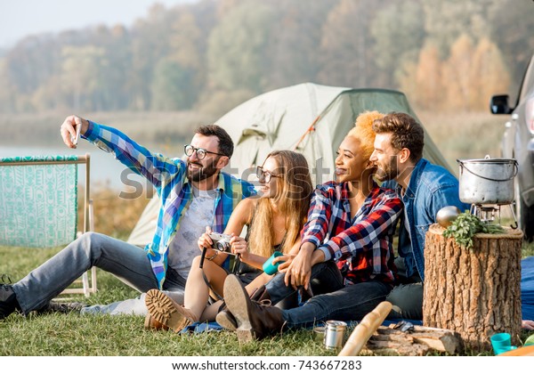 Multi ethnic group of\
friends dressed casually having fun making a selfie photo together\
during the outdoor recreation with tent, car and hiking equipment\
near the lake