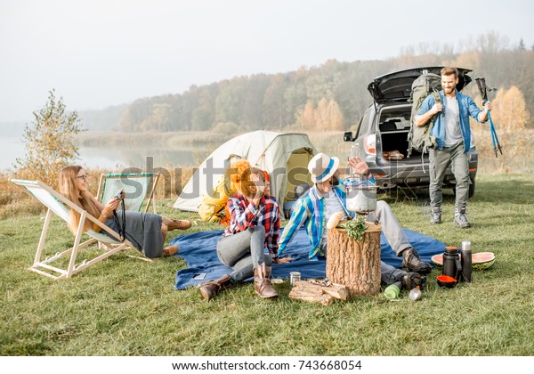 Multi ethnic group of friends dressed casually\
having a picnic during the outdoor recreation with tent, car and\
hiking equipment near the\
lake