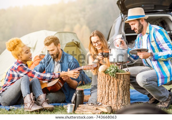 Multi ethnic group of friends dressed\
casually having a picnic, cooking soup with cauldron, playing\
guitar during the outdoor recreation near the\
lake