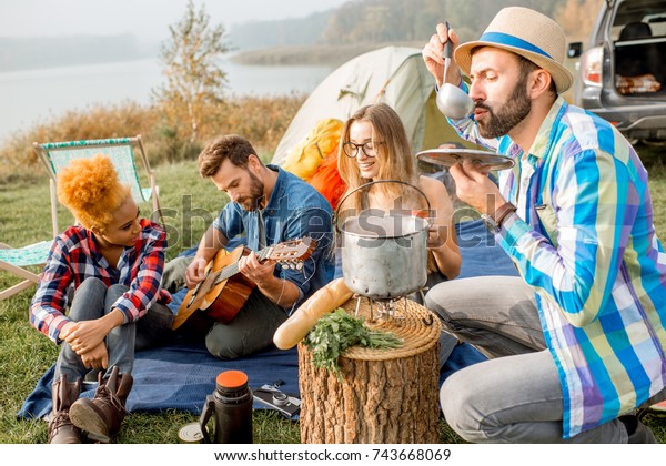 Multi ethnic group of\
friends dressed casually having a picnic, cooking soup with\
cauldron during the outdoor recreation with tent, car and hiking\
equipment near the lake