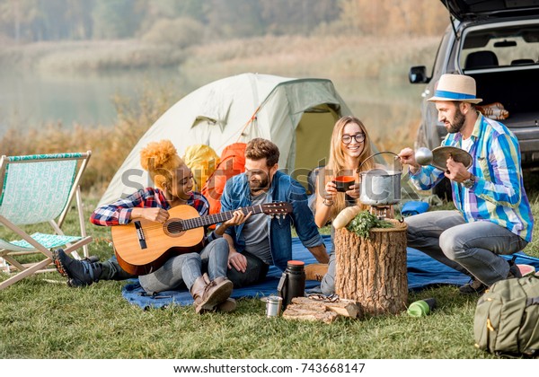 Multi ethnic group of friends dressed\
casually having a picnic, cooking soup with cauldron, playing\
guitar during the outdoor recreation near the\
lake