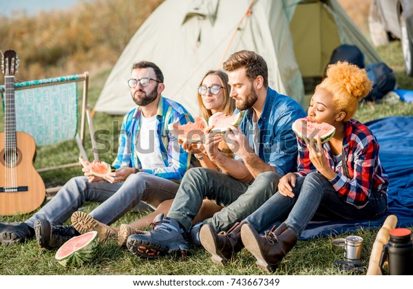 Multi ethnic group of friends having a picnic,\
eating watermelon during the outdoor recreation with tent, car and\
hiking equipment near the\
lake