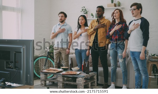 Multi ethnic group of friends sport fans singing\
national anthem before watching sports championship on TV together\
at home indoors
