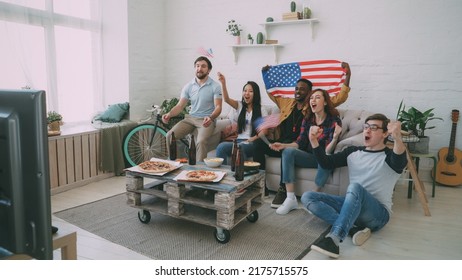 Multi Ethnic Group Of Friends With USA National Flags Watching Sport Championship On TV Together Cheering Up Their Favourite Team At Home Indoors