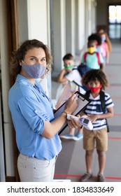 Multi Ethnic Group Of School Children Wearing Face Masks, Standing In Line, Having Their Temperature Checked By A Teacher. Education Back To School Health Safety During Covid19 Coronavirus Pandemic.