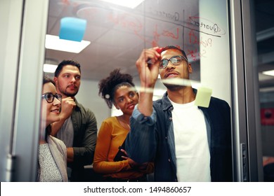 Multi ethnic happy creative business team looking at young man writing ideas on glass board with red marker at office - Powered by Shutterstock