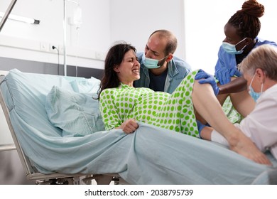 Multi Ethnic Medical Team Delivering Child For Woman In Labor Laying In Bed At Maternity. Young Adult With Contractions Giving Birth To Baby In Hospital Ward With Husband, Doctor And Nurse