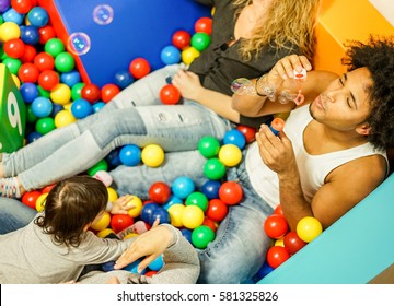 Multi Ethnic Parents Playing With Daughter Inside Ball Pit Swimming Pool - Happy People Having Fun In Children Playground Indoor - Family And Love Concept - Focus On Man Face - Warm Filter