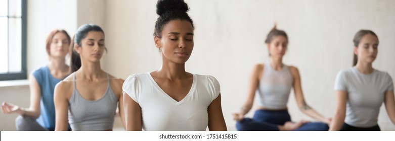 Multi Ethnic Pretty Women Seated Cross-legged Do Meditation Practice During Group Work Out Yoga Session At Sport Club Lead By African Female Trainer. Horizontal Photo Banner For Website Header Design