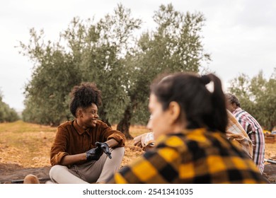 Multi ethnic team of farmers are taking a break and chatting together during the olive harvest in a sunny orchard, wearing gloves and casual work clothes - Powered by Shutterstock