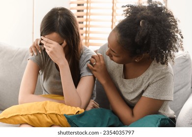 Multi ethnic young African woman friend consoling her crying Asian ethnic friend while sitting on sofa. People mental health care friendship diversity people relation concept. - Powered by Shutterstock