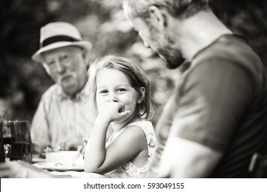 Multi Generation Family Gathered To Lunch In The Garden In Summer, Focus On A Beautiful Little Girl Looking At Camera. Black And White