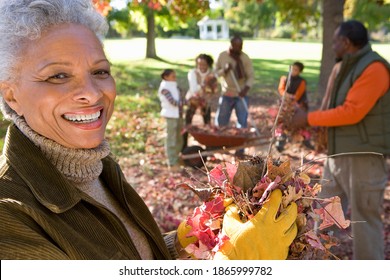 Multi Generational Family Gathering Autumn Leaves In A Garden With Focus On The Grandmother In The Foreground Smiling At The Camera.