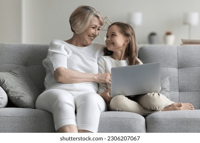 Multi generational family, grandma and granddaughter sit on couch with wireless computer, look at each other with tenderness and love, enjoy leisure on internet and time together, watch on-line movies
