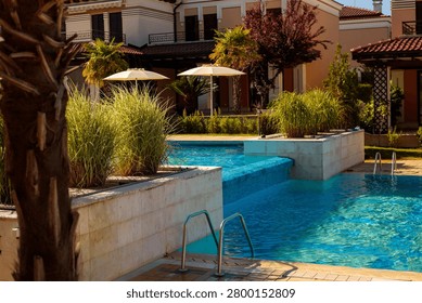 A multi level swimming pool features a tiled waterfall edge connecting an upper section to the main pool, framed by a tropical garden of ornamental grasses and palm trees. Two white closed umbrellas stand near a beige villa building with a red tiled roof and decorative balconies. Metal handrails mark the pool entry steps on the right side. This warmly lit resort pool scene combines architectural elegance with lush tropical landscaping for an inviting outdoor retreat