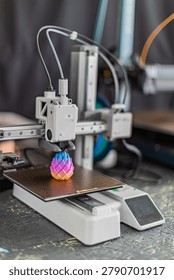 Multiaxis 3D printer with metallic frame, white extruder head depositing material onto dark build plate, showcasing gradient color scheme from blue to orange, textured surface design, blurred backgr.