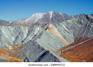 Multicolor autumn landscape with snow-covered mountain and gray rockies with orange and lilac tint. Spectacular colorful view to sharp mountain ridge in fall. Motley mountain scenery in autumn colors.