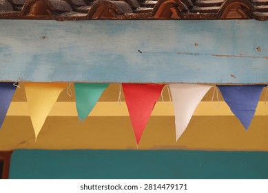 Multicolor triangular flags hanging on a wooden house beam as party or festival decoration. Row of colorful triangular bunting flags hanging under the wooden eaves of a traditional clay tile roof.