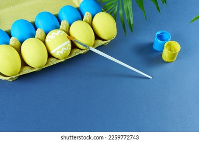 Multicolored Easter eggs packaged on a blue background with a brush and cans of paint. The concept is preparation for the holiday.