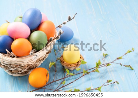 Multi-colored Easter eggs in a wicker basket on a blue wooden table with twigs of birch with young leaves - Easter composition with copy space