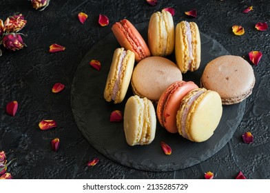 Multicolored Macaroons On Slate, On A Black Concrete Background With Red Rose Petals. Selective Focus With Copy Space. View From A High Angle