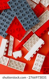 Multicolored plastic building blocks create a dense and dynamic texture against a bright red background. The chaotic accumulation of white, gray, and red pieces symbolizes creative mess, limitless children's imagination,