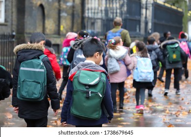 Multi-Cultural Children (asian, Indian, Chinese, Caucasian) primary student or kids and teacher carrying  school bag walk in street in London in rain winter day, with maple leaves on ground. back view