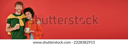 multicultural couple holding cups of coffee on coral background, morning routine, cultural diversity, vibrant colors, stylish outfits, interracial people holding mugs and looking at camera, banner