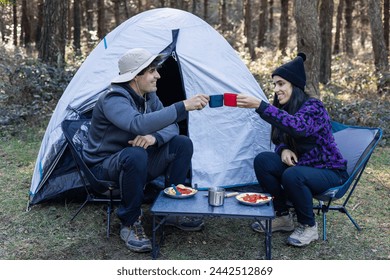 multicultural couple from the previous image is now smiling and joyfully toasting with their mugs at the campsite, sharing a moment of happiness and connection during their outdoor adventure - Powered by Shutterstock