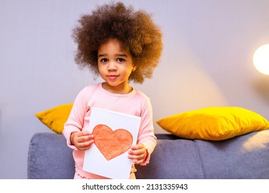 Multicultural Cute Little Curly Girl Holding Post Card With Drawn Heart In Cozy Living Room At Evening