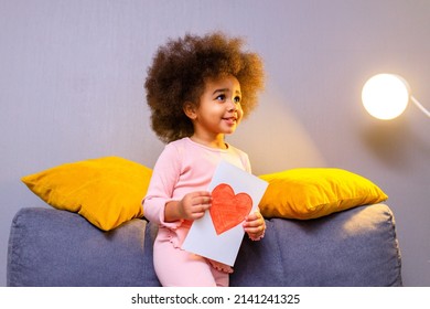 Multicultural Cute Little Curly Girl Holding Post Card With Drawn Heart In Cozy Living Room At Evening