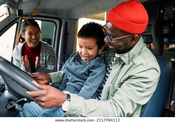 A multicultural father holds\
his toddler son in a lap and drives their home on the wheels while\
a mother applauds and looks at them with a smile on her\
face.