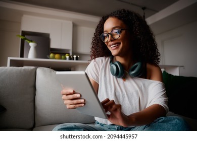 Multi-cultural female with glasses happily typing on tablet while sitting on the sofa, content