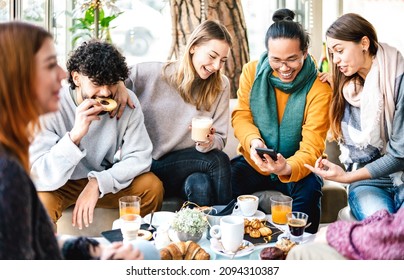 Multicultural friends playing with mobile phone at coffee bar - People having fun together at cafeteria on brunch time - Life style concept with happy men and women at cafe venue - Bright warm filter 