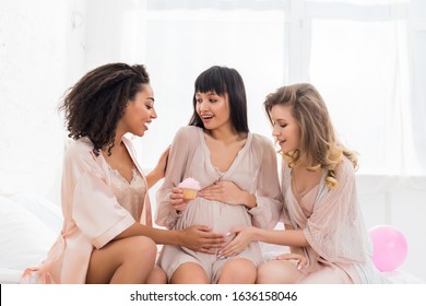 multicultural girlfriends touching tummy of young pregnant woman holding pink cupcake on baby shower  - Powered by Shutterstock