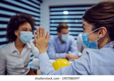 Multicultural Group Of Architects With Face Masks Sitting In Boardroom And Having Meeting About New Project. Selective Focus On Woman Giving High Five To Her Female Colleague.