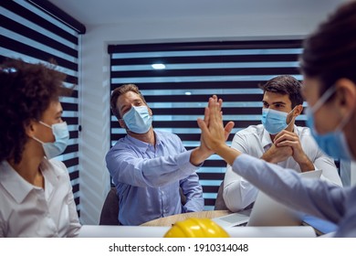Multicultural Group Of Architects Sitting In Office With Face Masks And Having Meeting About New Project During Corona Virus. Selective Focus On Man Giving High Five To His Female Colleague.