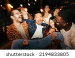A multicultural group of friends are lifting their male tipsy friend at the rooftop night outdoor party.