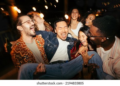 A multicultural group of friends are lifting their male tipsy friend at the rooftop night outdoor party. - Shutterstock ID 2194833805