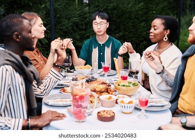 Multicultural group of people celebrating Thanksgiving together and holding hands sitting at dinner table outdoors shot with flash - Powered by Shutterstock