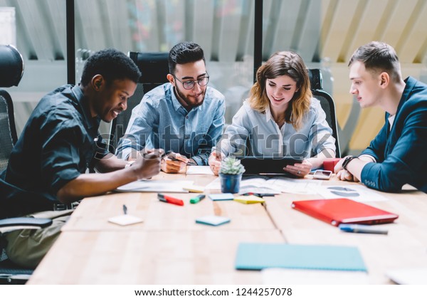 Mångkulturell grupp av professionella designers som samarbetar på uppgiften med hjälp av modern pekplatta enhet under brainstorming möte i kontorsinredning. Fyra anställda diskuterar arbete med produktivitet