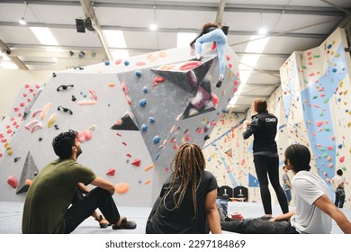 Multi-cultural group sitting and watching friend on climbing wall in indoor activity centre being taught by coach - Powered by Shutterstock