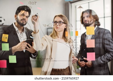 Multicultural Group Of Young Financiers In Formal Wear Using Transparent Glass Board During Office Meeting. Female Worker Explaining New Strategy To Male Colleagues In Boardroom.