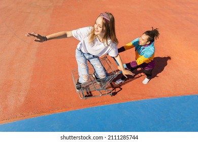 Multicultural group of young friends bonding outdoors and having fun - Stylish cool teens gathering at urban skate park, playful young couple doing cart race